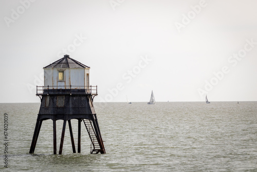 Fototapeta Naklejka Na Ścianę i Meble -  Old lightouse in the sea, Dovercourt low lighthouse, built in 1863 and discontinued in 1917 and restored in 1980 the 8 meter lighthouse is still a iconic sight, with sailing boats sailing past