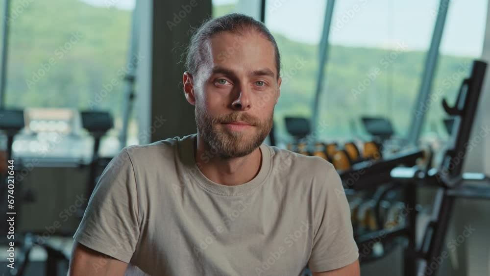 Close-up shot of young man in workout clothes sitting on bench and looking at camera. Good-looking bearded Caucasian male guy with gray eyes wearing t-shirt. Blurred gym background. Indoors