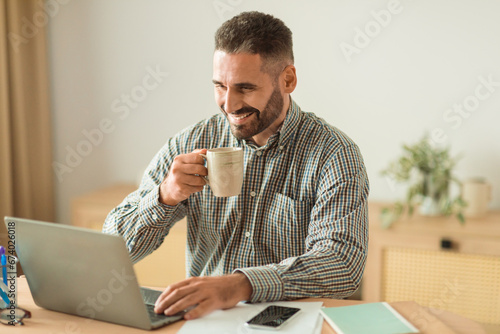 Happy businessman drinking coffee while using laptop at workplace indoor