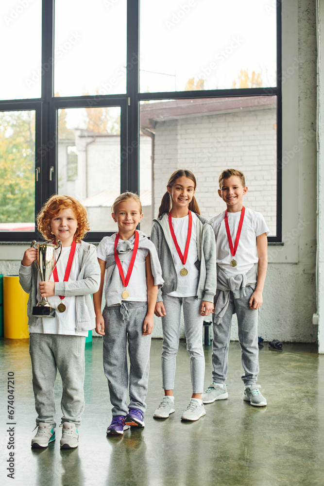 Fototapeta premium vertical shot of little boys and girls posing with golden medals and trophy, looking at camera