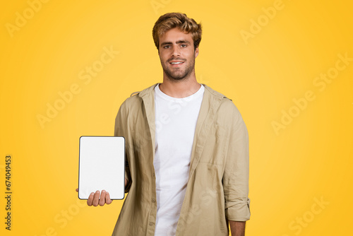 Joyful European man holding tablet with blank screen on yellow background