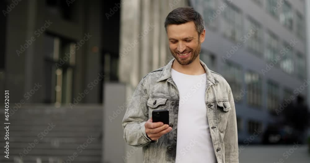 Smiling man wearing jeans jacket, leaving building, using smartphone.