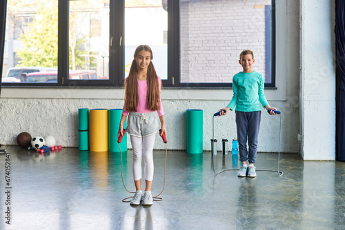 Photography little cute boy and girl in sportswear posing with jump ropes and smiling at cam
