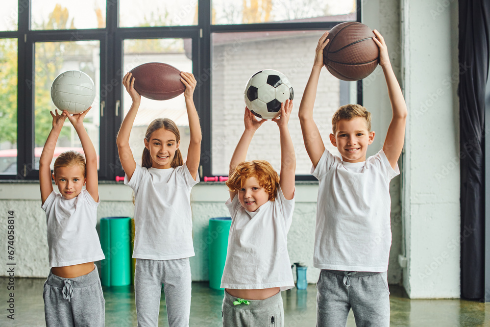 Fototapeta premium joyous little boys and girls posing with different types of balls above their heads, child sport