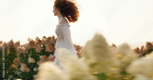 Beautiful red-haired woman in white elegant dress running on blooming field.