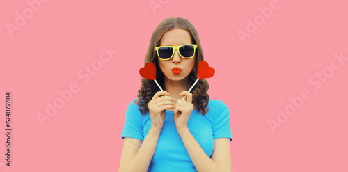 Portrait of beautiful young woman with red heart shaped lollipop blowing her lips with lipstick sending sweet air kiss wearing sunglasses on pink background