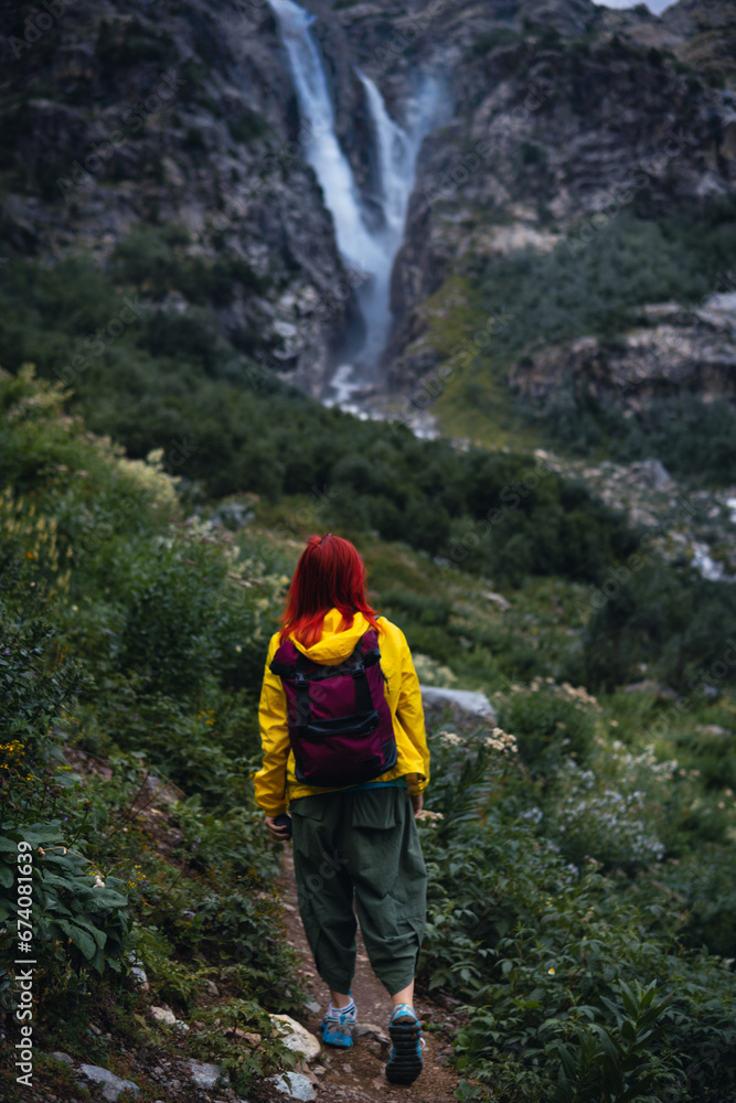 A woman, hiker walks along a path among high mountains. Shdugra waterfall in the background. Concept of hiking, active lifestyle, travel. Summer day, Georgia. Vertical photo