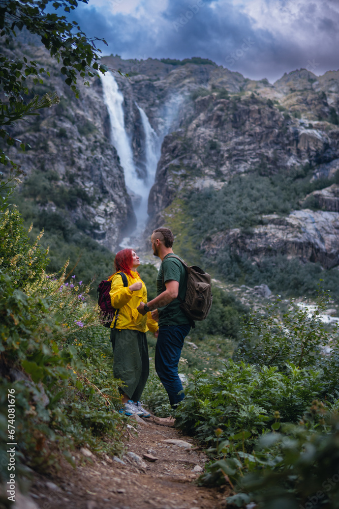 Beautiful young happy couple in love, woman and man with backpack. Shdugra waterfall in the background. Concept of hiking, active lifestyle, travel. Vertical photo. Summer day, Mazeri, Georgia Beautiful young happy couple in love, woman and man with backpack. Shdugra waterfall in the background. Concept of hiking, active lifestyle, travel. Vertical photo. Summer day, Mazeri, Georgia