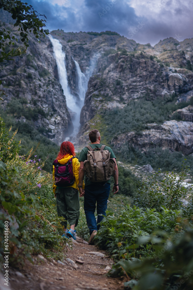 Back view of couple tourists, woman and man with backpack. Shdugra waterfall in the background. Concept of hiking, active lifestyle, travel. Vertical photo. Summer day, Mazeri, Georgia