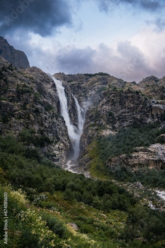 An incredible view of a majestic Shdugra waterfall, high in the Caucasus Mountains, rocky slopes, a rushing stream. Green herbs and plants with flowers. Mazeri, Svaneti, Georgia. Vertical photo