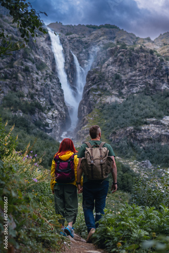 Back view of couple tourists, woman and man with backpack. Shdugra waterfall in the background. Concept of hiking, active lifestyle, travel. Vertical photo. Summer day, Mazeri, Georgia