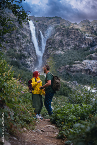 Beautiful young happy couple in love, woman and man with backpack. Shdugra waterfall in the background. Concept of hiking, active lifestyle, travel. Vertical photo. Summer day, Mazeri, Georgia