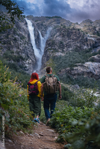 Back view of couple tourists, woman and man with backpack. Shdugra waterfall in the background. Concept of hiking, active lifestyle, travel. Vertical photo. Summer day, Mazeri, Georgia