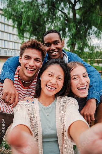 Vertical portrait of a group of young multiracial high school students with a toothy smile taking a selfie outside looking at camera, laughing and having fun together. Friendly teens shooting a photo