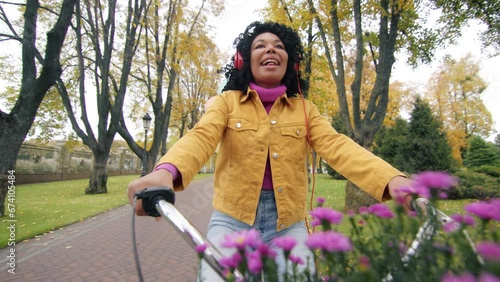 Closeup view of dark-haired curly Afro-American lady in headphones on a bike with chrysanthemums and balloons, singing, enjoying the view of autumn park. High quality 4k footage photo