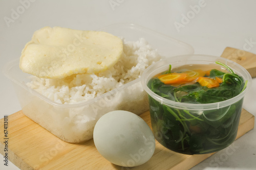 a menu of white rice with clear vegetables of spinach leaves and carrots, side dishes of shrimp crackers and a salted egg on a wooden board isolated on a white background