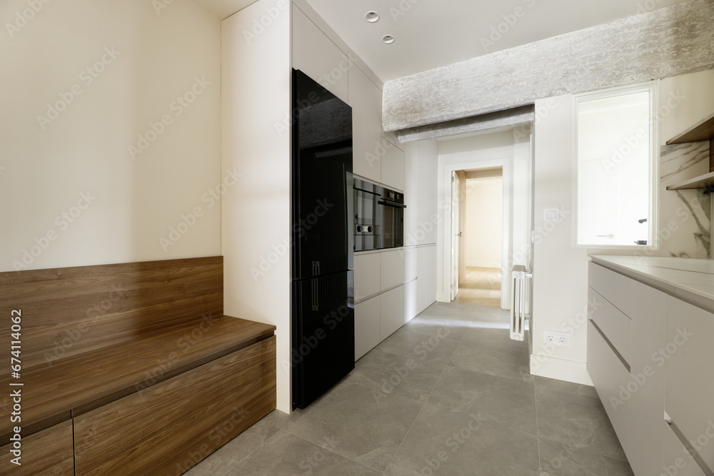 Newly installed narrow kitchen with white handleless furniture on one side and a long wooden bench with cabinets on the other, integrated appliances with black glass doors and gray stone floors Newly installed narrow kitchen with white handleless furniture on one side and a long wooden bench with cabinets on the other, integrated appliances with black glass doors and gray stone floors