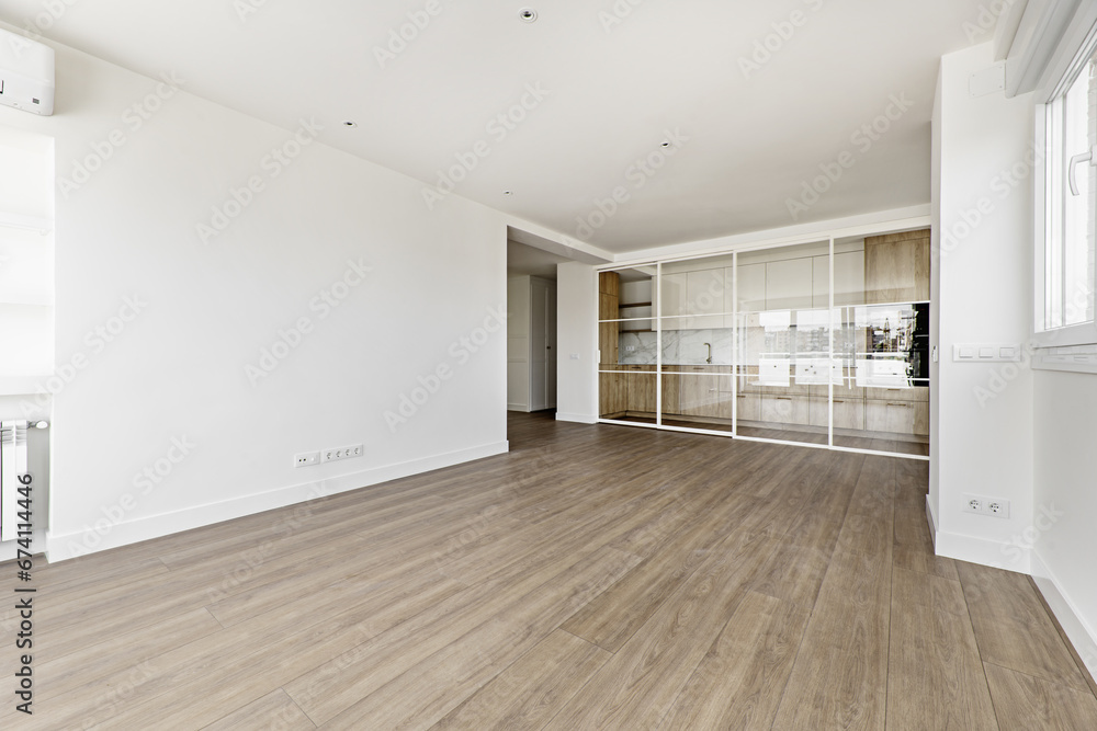 Fototapeta premium Large empty modern living room with white painted walls and sliding glass partition enclosing a kitchen with wooden furniture
