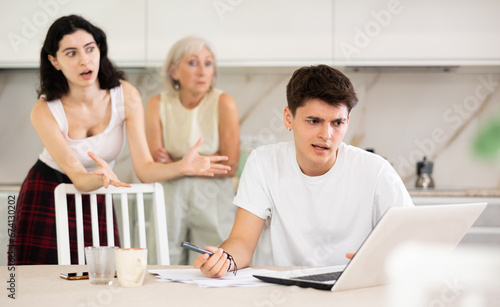 Young guy while working on laptop listens to claims of elderly mother and young sister girl in incompetence, unsuccessful study results