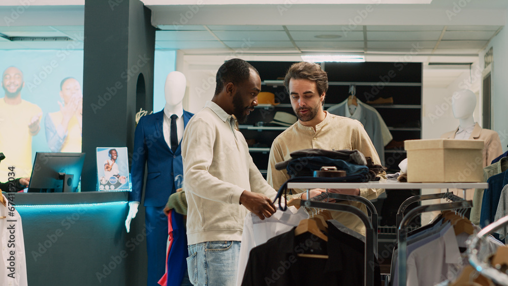 African american man asking assistant for help with shirts, looking to buy formal wear on hangers. Retail store employee talking to client about shopping for fashionable clothes. Tripod shot. African american man asking assistant for help with shirts, looking to buy formal wear on hangers. Retail store employee talking to client about shopping for fashionable clothes. Tripod shot.