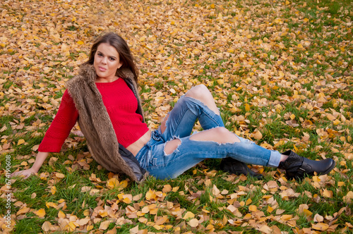 Sexy and attractive young woman in red and torn jeans is sitting on the ground covered with fallen leaves