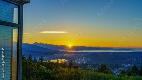 Sunrise over distant mountains in silhouette shining through early morning mist over Fraser Valley and Port Moody, BC, as seen from a Burnaby Mountain balcony.