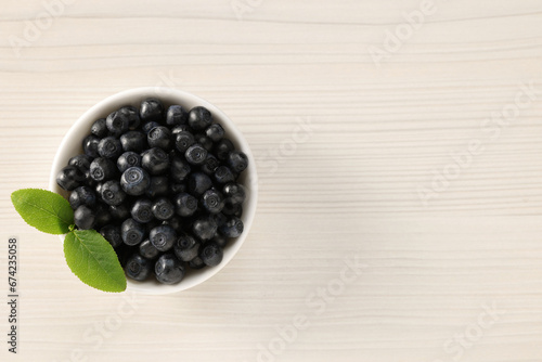 Tasty fresh bilberries with leaves in bowl on light wooden table, top view. Space for text