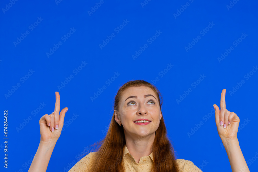 Redhead woman showing thumbs up and pointing empty place over head, advertising area for commercial text, copy space for goods promotion advertisement. Adult girl isolated on blue studio background Redhead woman showing thumbs up and pointing empty place over head, advertising area for commercial text, copy space for goods promotion advertisement. Adult girl isolated on blue studio background