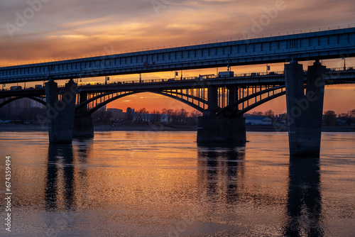 Beautiful view of the bridge over which cars drive at sunset. A river flows under the bridge, reflecting the sunset rays. Bridge in the city of Novosibirsk, Russia © Анатолий Савицкий