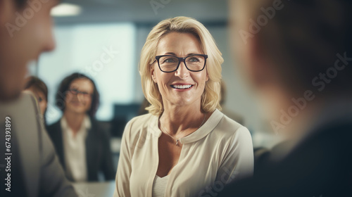 Smiling mature businesswoman in eyeglasses looking at camera during meeting with colleagues in office. Generative Ai. 