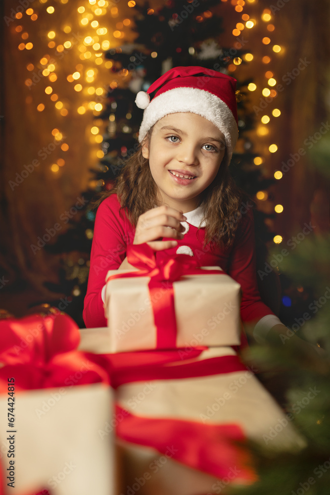 Happy 6 years old girl in Santa Claus costume wraps gifts boxes with red ribbons and festive lights on background Happy 6 years old girl in Santa Claus costume wraps gifts boxes with red ribbons and festive lights on background