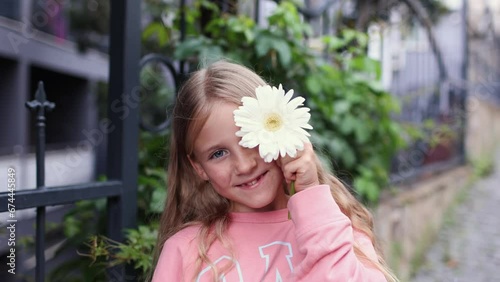 Cute little blonde girl holds big white flower near one eye and smiles to camera, adorable little girl with white gerbera at the street