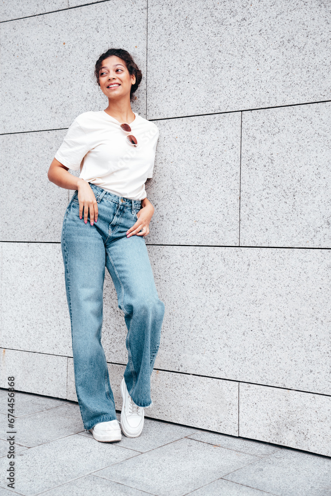 Young beautiful smiling hipster woman in trendy summer white t-shirt and jeans clothes. Carefree woman, posing in the street at sunny day. Positive model outdoors near wall Young beautiful smiling hipster woman in trendy summer white t-shirt and jeans clothes. Carefree woman, posing in the street at sunny day. Positive model outdoors near wall