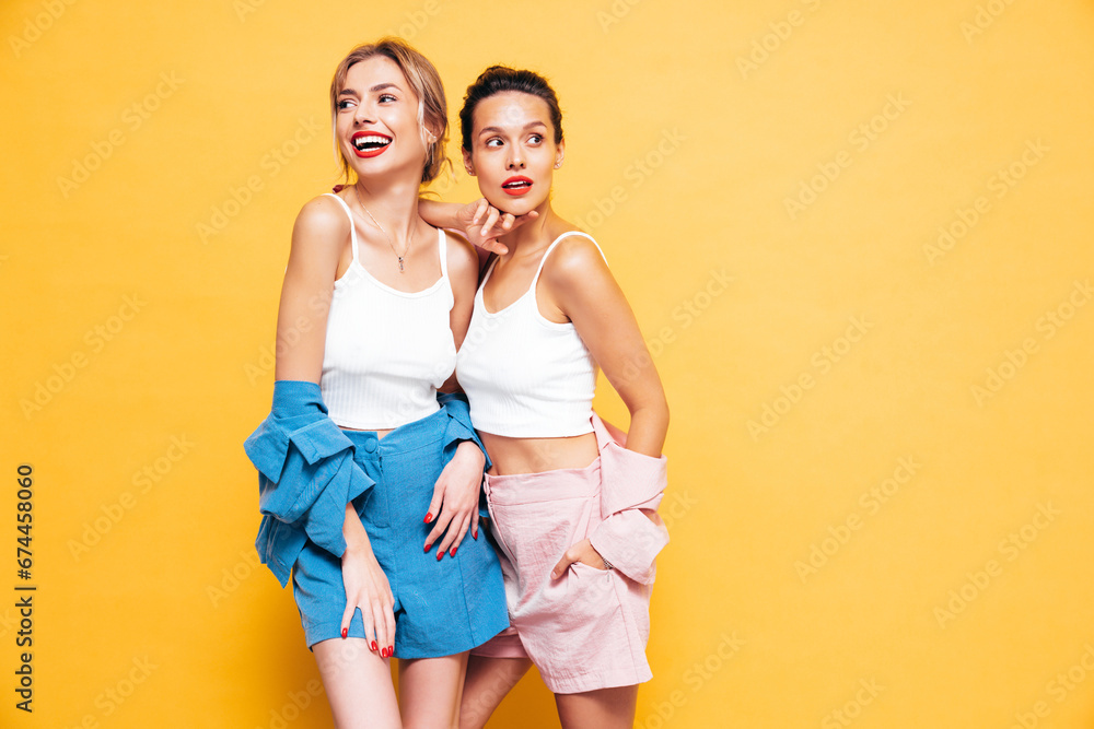 Two young beautiful smiling brunette hipster female in trendy summer clothes. Sexy carefree women posing near yellow wall in studio. Positive models having fun. Cheerful and happy Two young beautiful smiling brunette hipster female in trendy summer clothes. Sexy carefree women posing near yellow wall in studio. Positive models having fun. Cheerful and happy