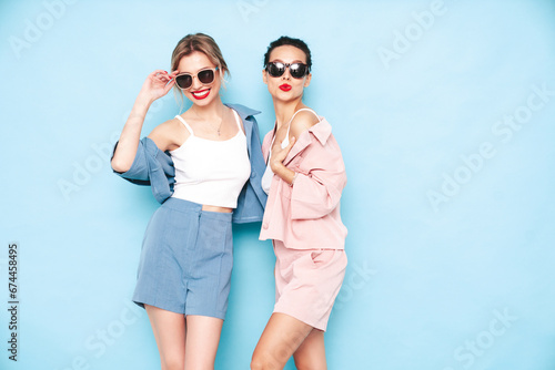 Two young beautiful smiling brunette hipster female in trendy summer clothes. Sexy carefree women posing near blue wall in studio. Positive models having fun. Cheerful and happy © halayalex