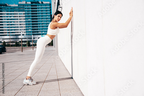Fitness woman in sports clothing. Sexy young beautiful model athlete doing fitness workout. Female making exercises in the street at sunny day. Stretching out before training. near wall