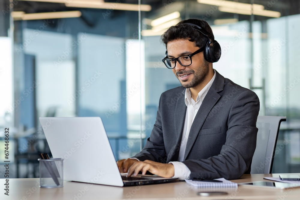 Successful indian programmer working inside office with laptop, man in headphones and business suit, typing code on keyboard, developing new software.