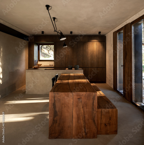 Front view modern wooden kitchen with a large rough concrete island and a large wooden table, two black chairs and a bench thought to be made of wood. photo