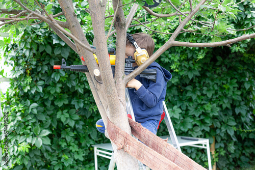 The boy looks into the optical sight of a children's water weapon. The child made a sniper point on the tree. Children play with a water pistol outdoors. A war simulator game.
