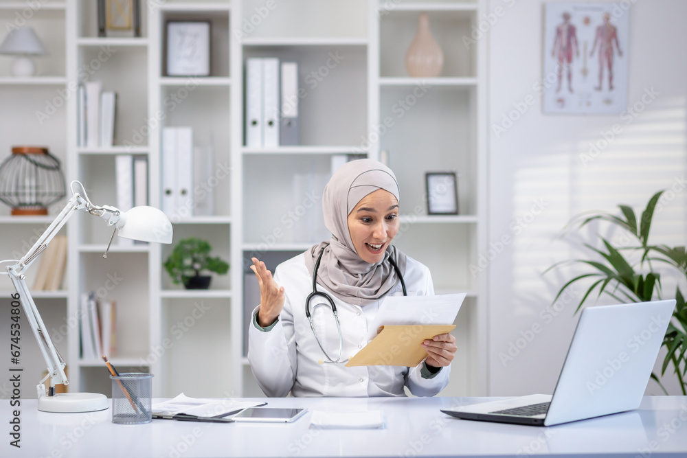 Joyful female doctor in hijab received envelope message mail notification with good results of patient's treatment, woman is happy and smiling, working inside clinic office in white medical coat. Joyful female doctor in hijab received envelope message mail notification with good results of patient's treatment, woman is happy and smiling, working inside clinic office in white medical coat.