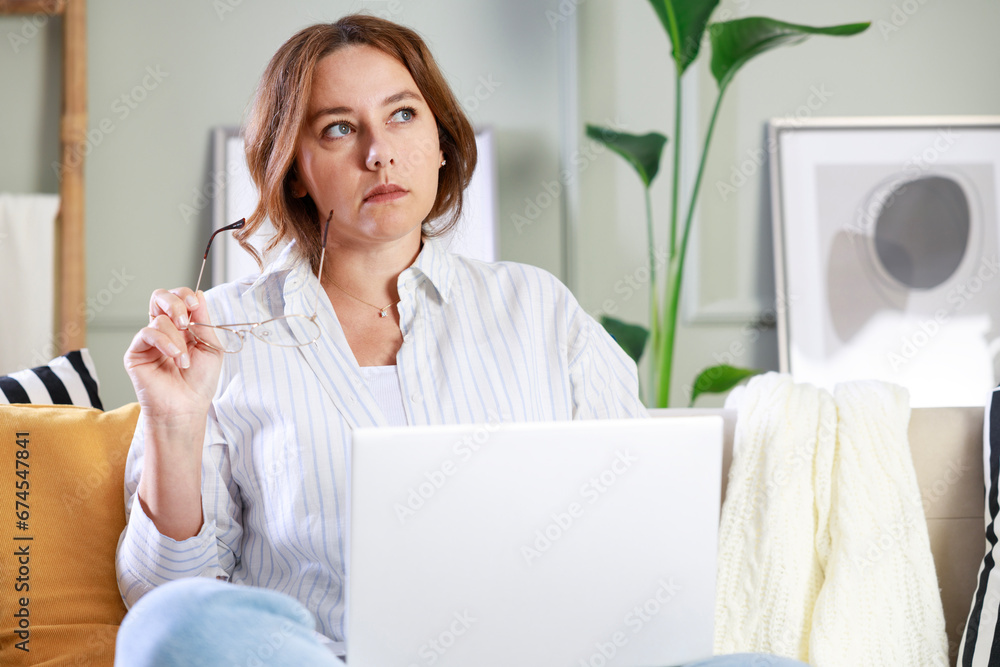 Beautiful young woman thinking while using laptop for blogging in living room at home