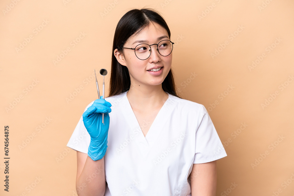 Dentist Chinese woman holding tools isolated on beige background looking to the side and smiling Dentist Chinese woman holding tools isolated on beige background looking to the side and smiling