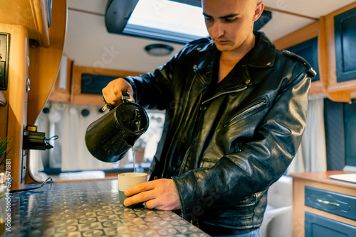 A man in the kitchen of a motorhome pours boiling water from a kettle into cup to make morning tea