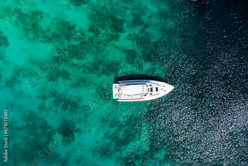 A yacht sails through crystal-clear turquoise waters, leaving a gentle wake behind. The aerial view captures the boat's sleek lines against the vibrant blue sea.