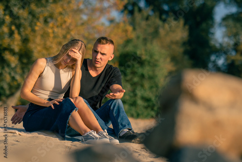 A young man quarrels with his girlfriend who turns away from him offended while sitting on sandy bank of the river photo