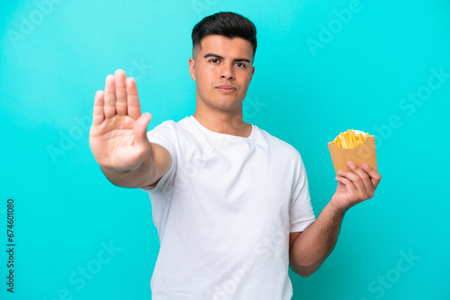 Young caucasian man catching french fries isolated on blue background making stop gesture © luismolinero