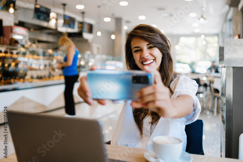 Beautiful and happy young woman sitting and eating delicious rolls in bakery or fast food. She also using her laptop and listening to music with headphones.