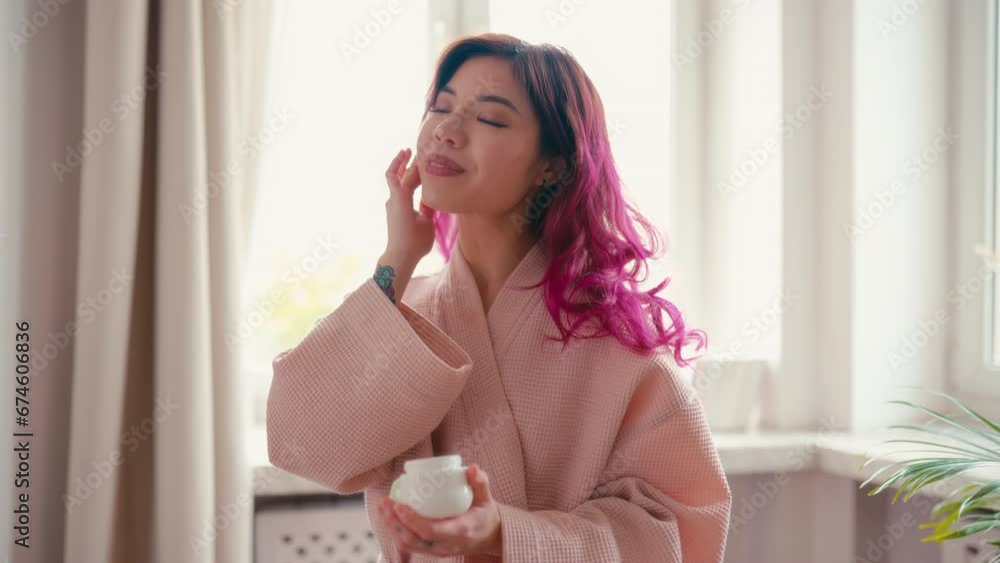 Beautiful young asian woman in bathrobe and pink hair applying moisturizing face cream after showering in bathroom. Spa treatments and face care.