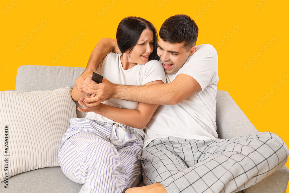 Young couple fighting for TV remote control while sitting on sofa against yellow background Young couple fighting for TV remote control while sitting on sofa against yellow background