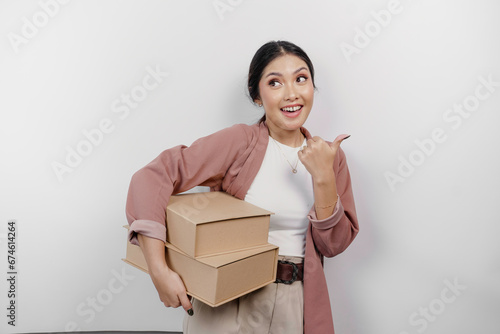 Excited young Asian woman employee is gesturing thumbs up for approval while carrying stack of cardboard boxes, isolated by white background © Reezky
